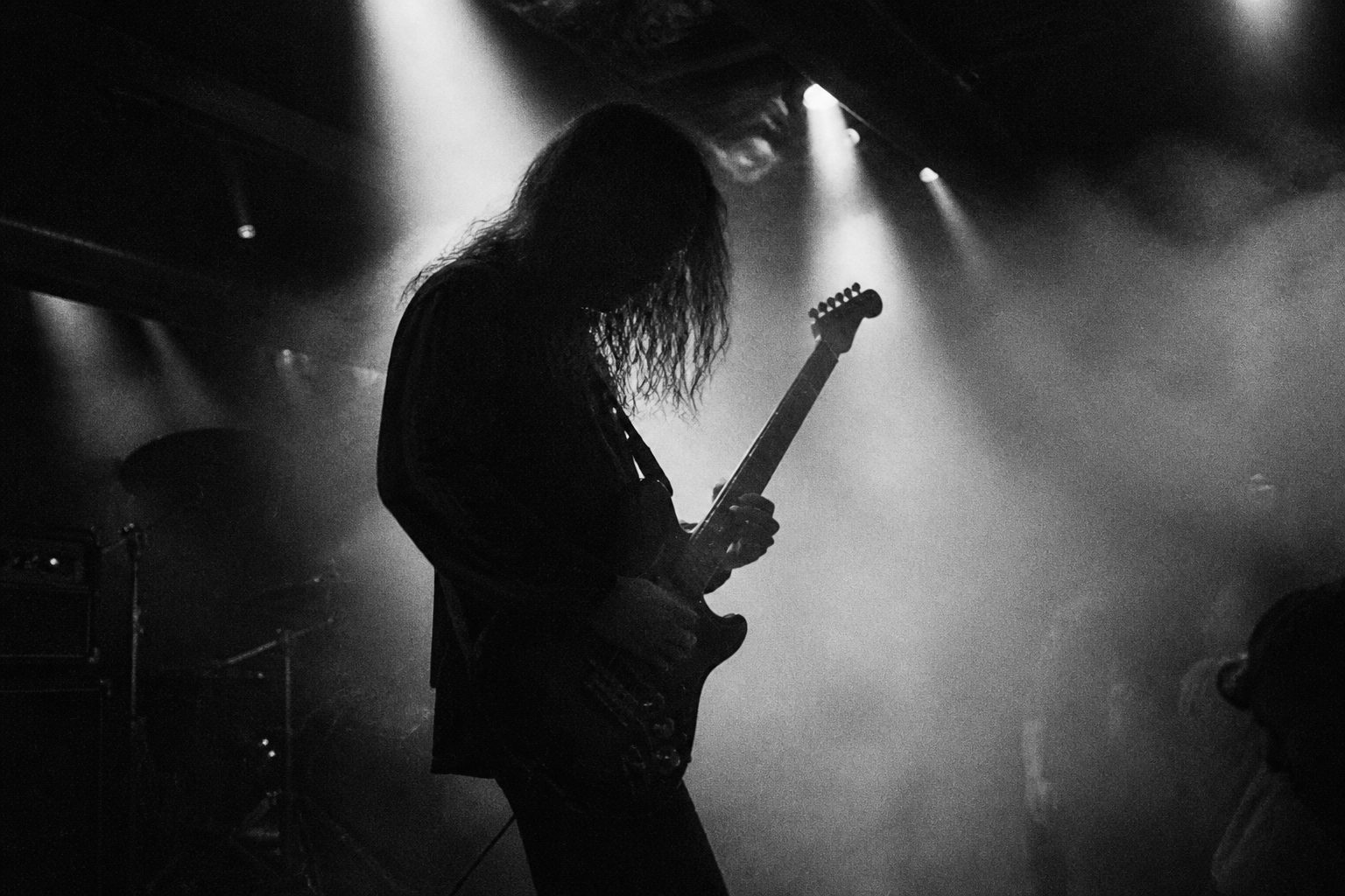 煙の中でギターを弾く長髪のギタリストの横顔。天井の照明が細い筋になって落ちる